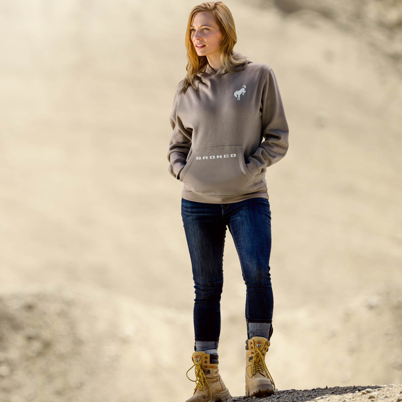 women standing in dirt scene with blue jeans and boots wearing a brown hoodie with bronco on pocket and horse on left shoulder