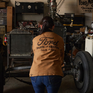 A person viewed from behind wearing a tan Ford Parts of Service jacket in a workshop, standing next to a vintage car frame with a large tire.