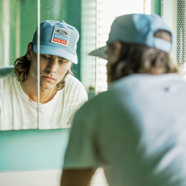 A man wearing a light blue Ford Performance cap and a white t-shirt looks at his reflection in a mirror, with the back of his head in the foreground.