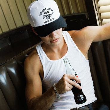 A man wearing a white and black trucker hat with 'Ford Motor Company' printed on it, sitting in a dark leather booth and holding a glass bottle of cola.