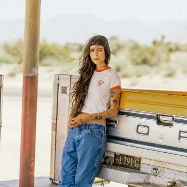 A woman with long dark hair leans against the back of a vintage Ford pickup truck in a desert setting. She is wearing a white short-sleeve ringer T-shirt with orange trim and a small red graphic on the chest, paired with blue jeans.