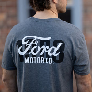 A man viewed from behind wearing a heather dark gray t-shirt with a large white and black Ford Motor Co. graphic on the back, set against a blurred brick wall background.