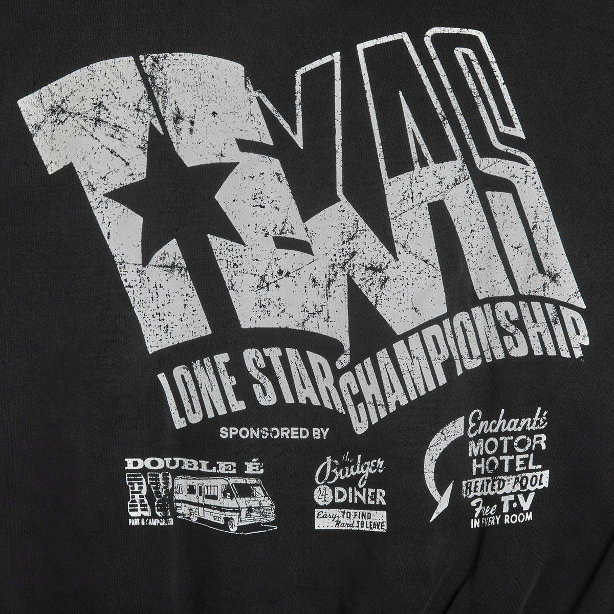Close-up of a black graphic T-shirt featuring a distressed white 'TNR' logo with a star, the words 'LONE STAR CHAMPIONSHIP,' and smaller sponsor-style prints including 'Double E,' 'Cruiser Diner,' and 'Enchanted Motor Hotel.'.