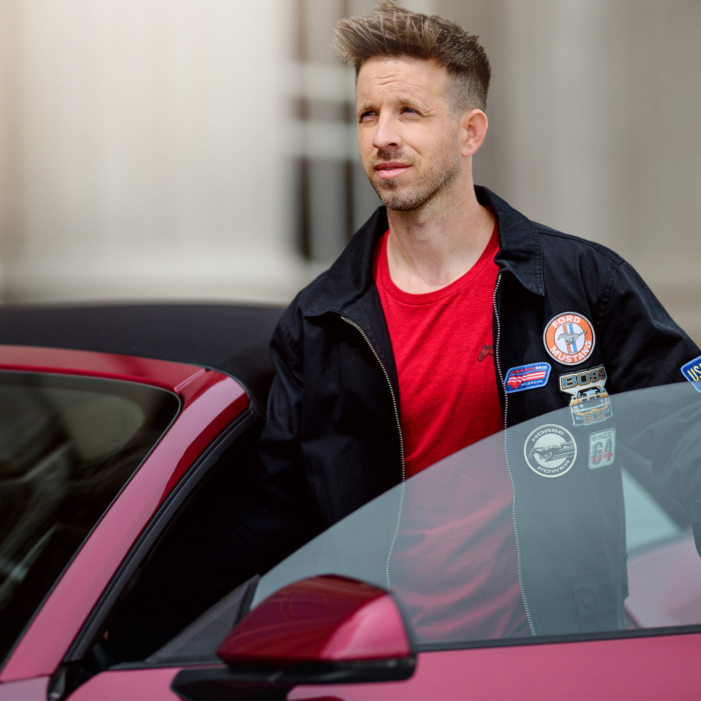 Man leaning against a red mustang car with a blurred background