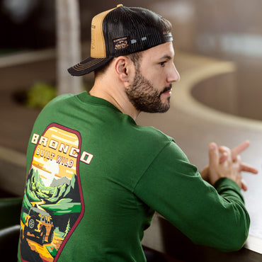 Man wearing a green sweatshirt with 'Bronco Built Wild' design, sitting indoors.