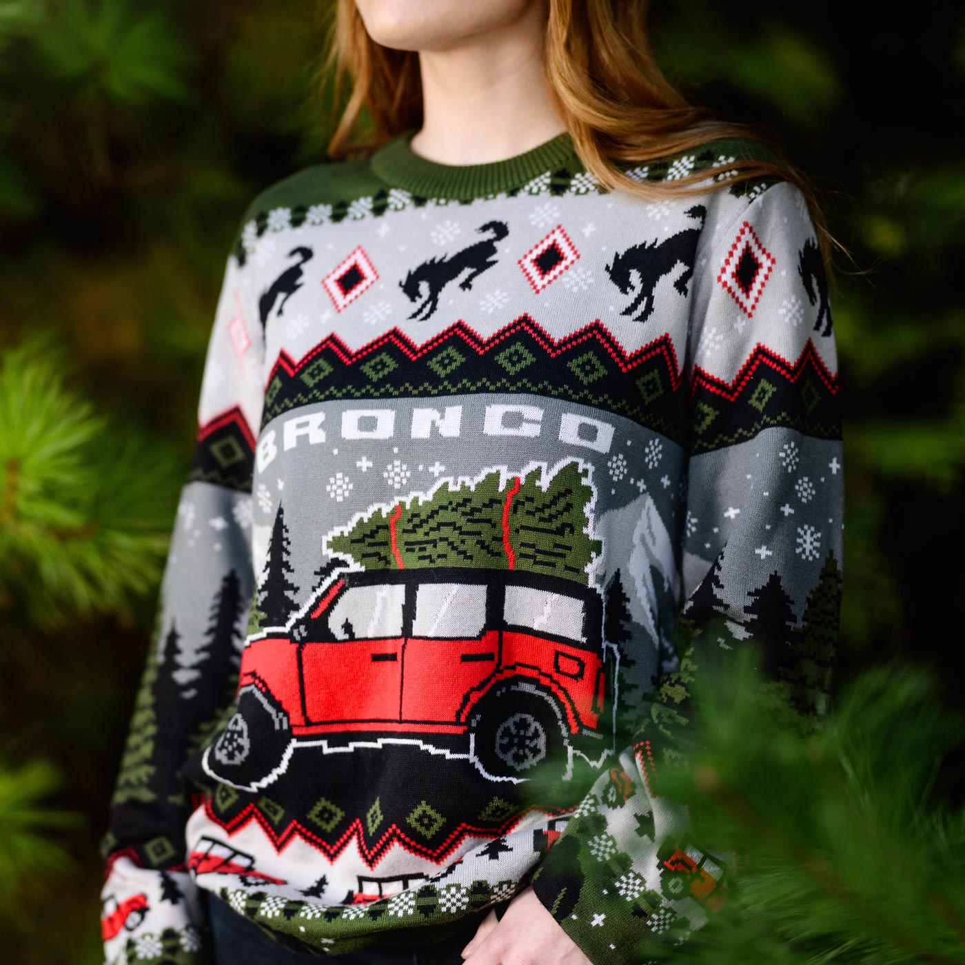 Person wearing a Christmas sweater with a red Bronco and tree design, standing among greenery.