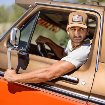 man hanging out door of vintage bronco truck wearing a tan bronco hat with a patch