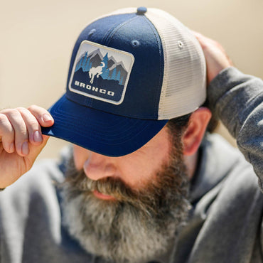 man with beard wearing navy blue and white bronco hat with a mountain scene bronco patch