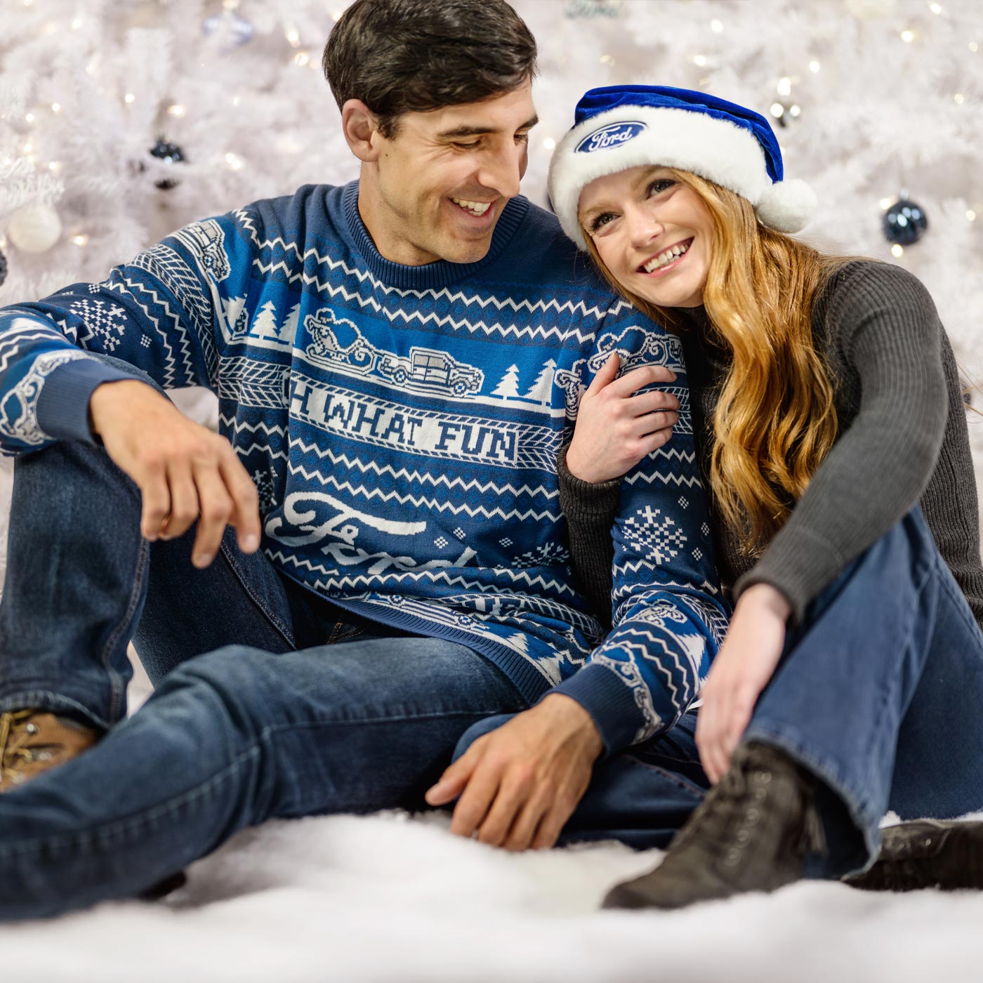 Man and woman sitting together wearing festive sweaters and a Ford hat against a snowy background.