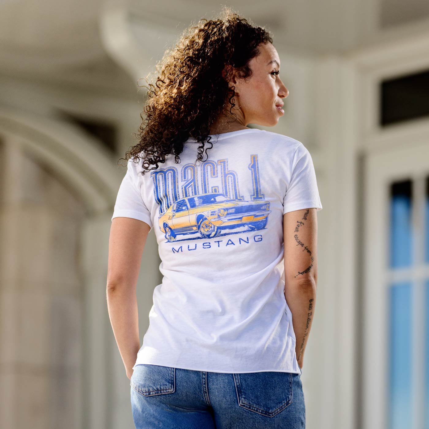 Woman wearing a white t-shirt with a graphic design of a mach 1 mustang classic car and text, standing in an outdoor setting.