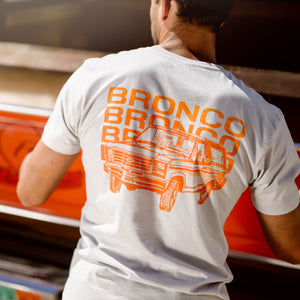 Person wearing a white t-shirt with orange 'Bronco' text and graphic, standing next to a red vehicle.