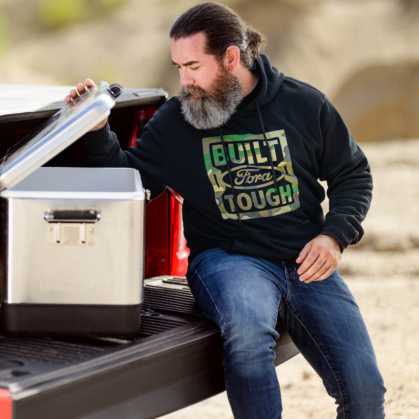 Man sitting on a truck bed with a cooler, wearing a hoodie with 'Built Ford Tough' text.
