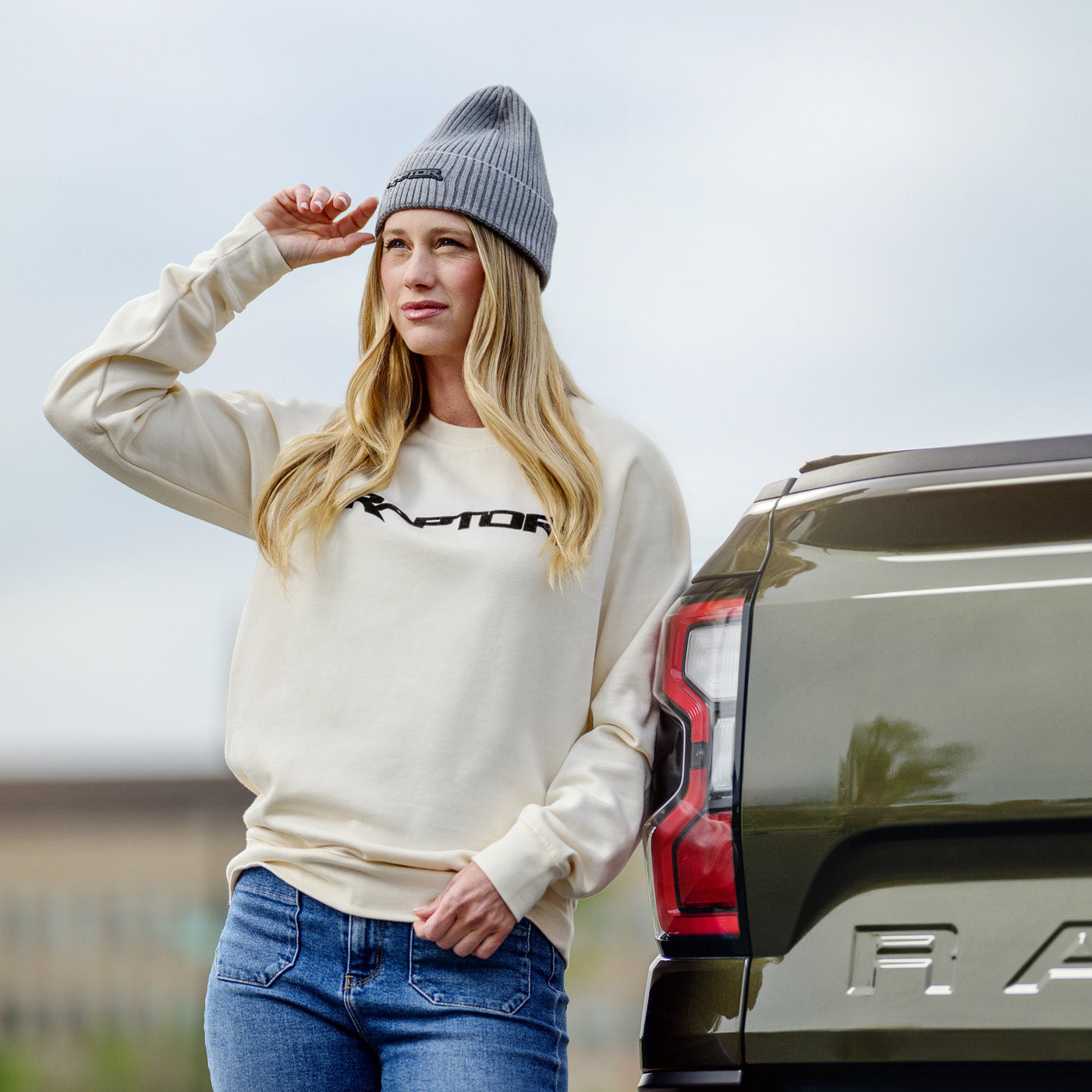 Woman wearing a cream sweatshirt with a logo, blue jeans, and a gray beanie standing next to a vehicle.