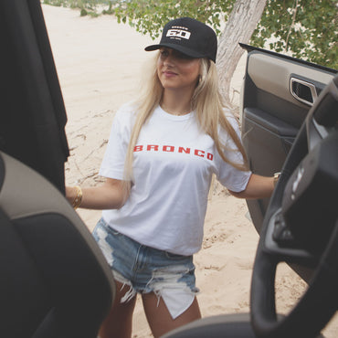 Woman wearing a 'Bronco' t-shirt and cap, standing next to an open car door outdoors.