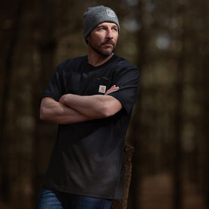 Man wearing a black t-shirt with a logo and a gray beanie standing in a forest.