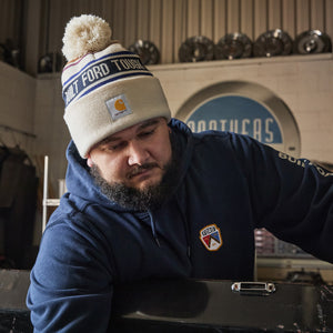 A man wearing a navy blue hoodie with a Carhartt logo patch and a beige beanie with a pom-pom and 'COLD FORD TOYS' text, leaning over a workbench in an automotive shop.
