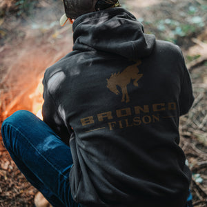 Person wearing a black hoodie with 'Bronco Filson' branding by a campfire in the woods.