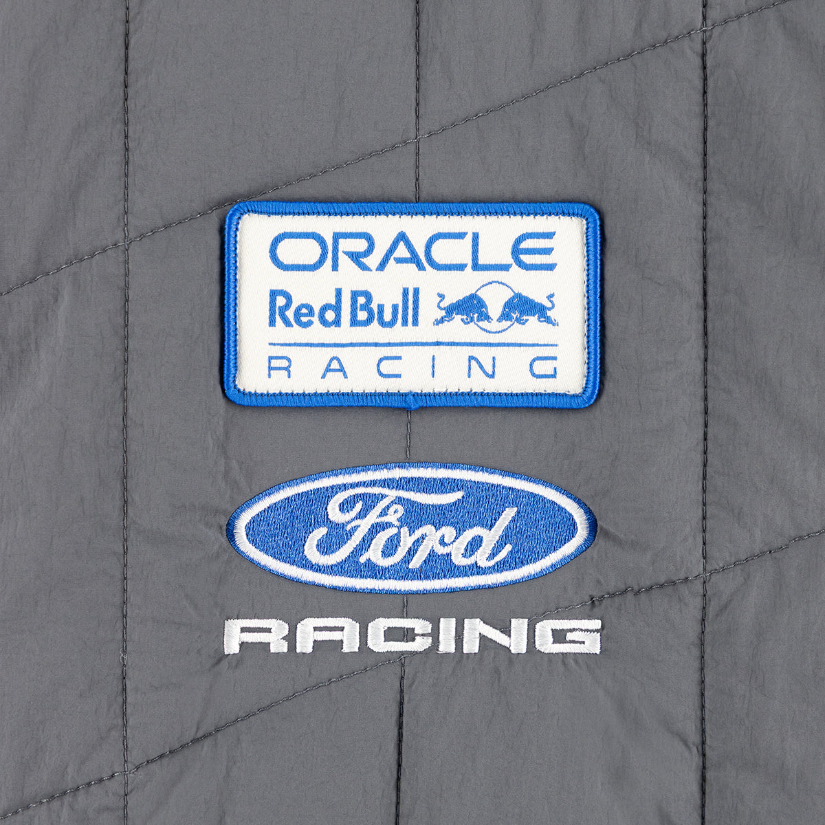 Close-up of a gray quilted racing jacket or pad featuring embroidered patches. The top patch reads 'ORACLE Red Bull RACING' in blue on white with a blue border, and the lower patch shows a blue Ford Racing oval logo above white 'RACING' text.