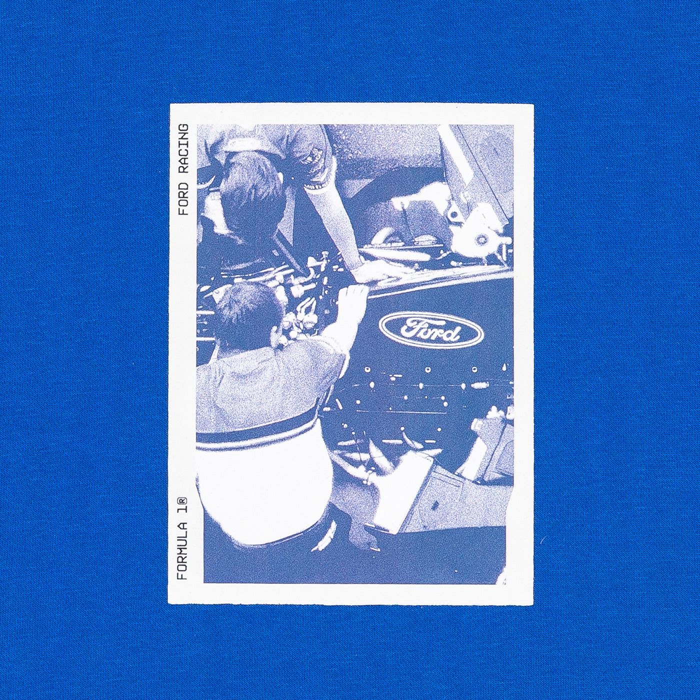 A vintage-style Ford racing collectible card featuring a blue-and-white photo of race drivers working around a car, displayed on a solid blue background.
