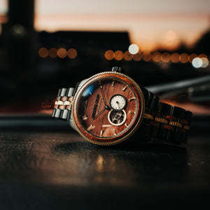 Wooden watch with metal band on a dark surface, blurred lights in the background