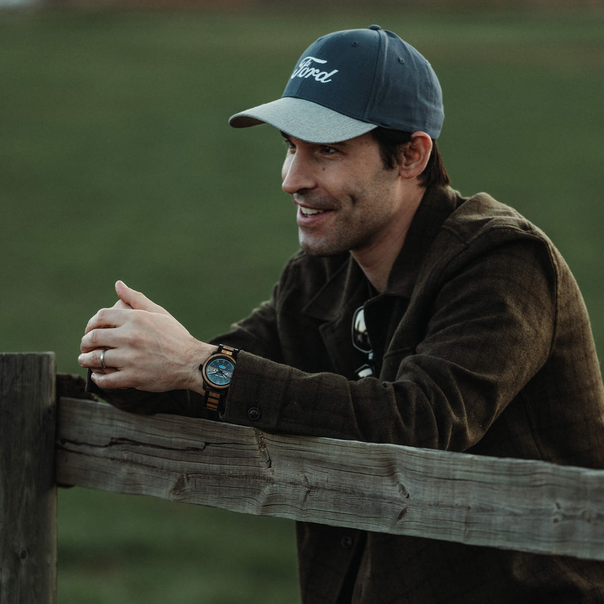 Man wearing a cap and jacket leaning against a wooden fence in a field
