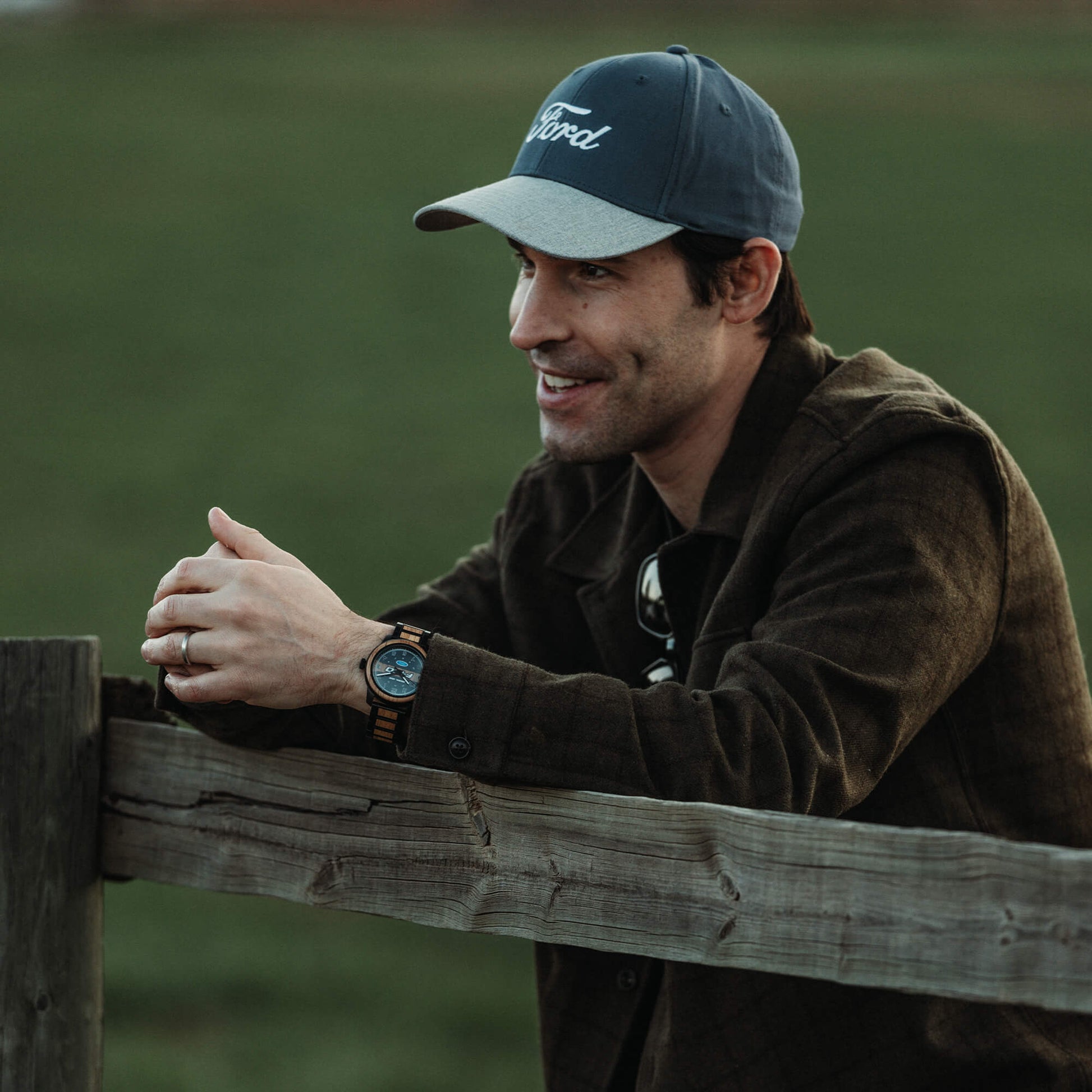 Man wearing a cap and jacket leaning against a wooden fence in a field