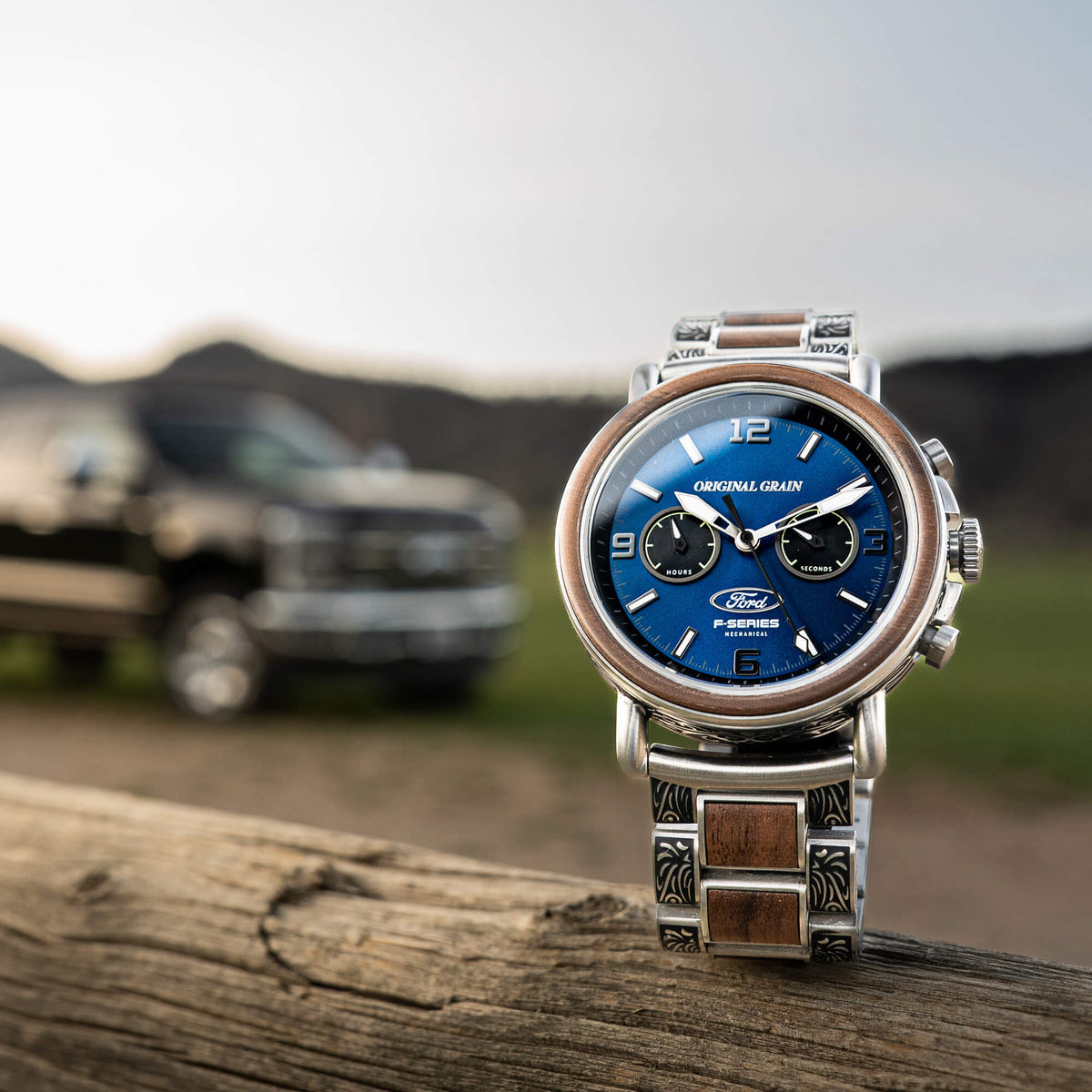 Silver watch with blue face on a wooden surface, blurred background of a truck and mountains
