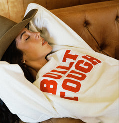 women in cowboy hat sleeping on a leather couch wearing a white built ford tough in red text crewneck sweatshirt from Ford x Brixton