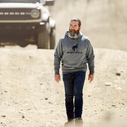 Man wearing a gray hoodie with 'Bronco' logo walking on a dirt road with a vehicle in the background.