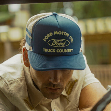 man in khaki jacket with eye covered by blue ford motor co truck country hat