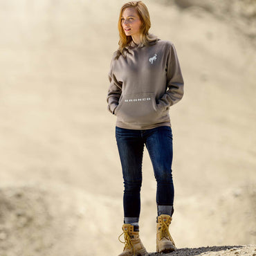 women standing in dirt scene with blue jeans and boots wearing a brown hoodie with bronco on pocket and horse on left shoulder
