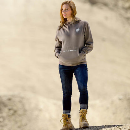 women standing in dirt scene with blue jeans and boots wearing a brown hoodie with bronco on pocket and horse on left shoulder