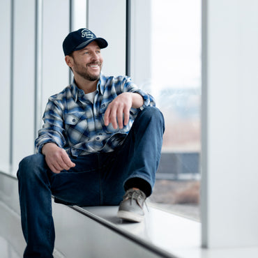 Man sitting on a windowsill wearing a plaid DIXXON shirt, cap, and jeans with a blurred outdoor background.
