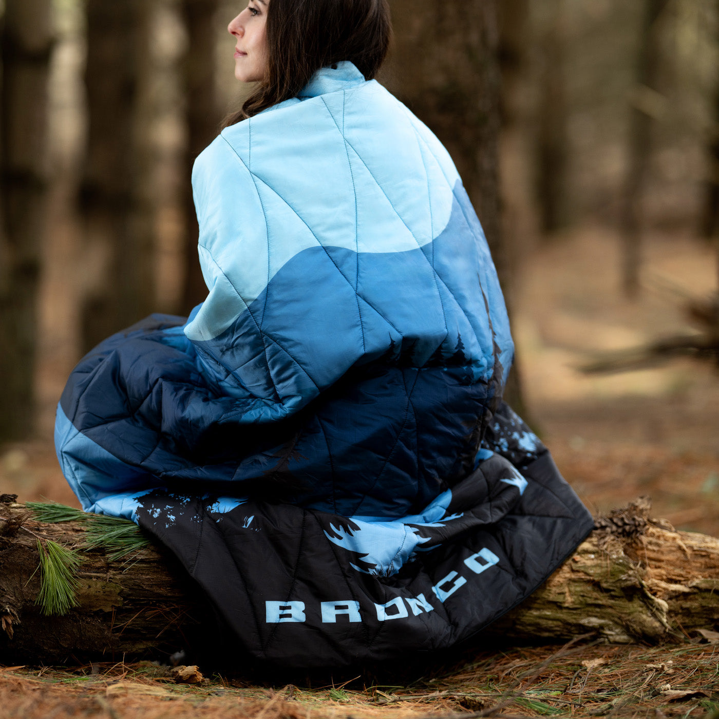 Person wrapped in a blue and black sleeping bag with 'Bronco' branding in a forest setting.