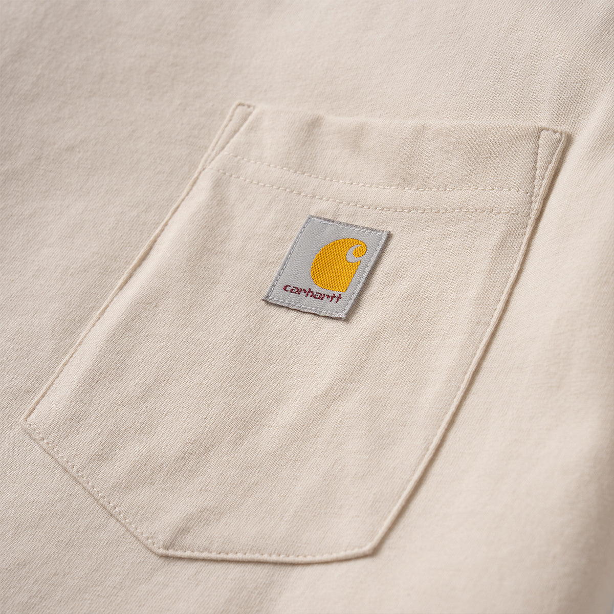 Close-up of a beige Carhartt pocket with the brand's signature yellow logo patch on a light gray fabric background.