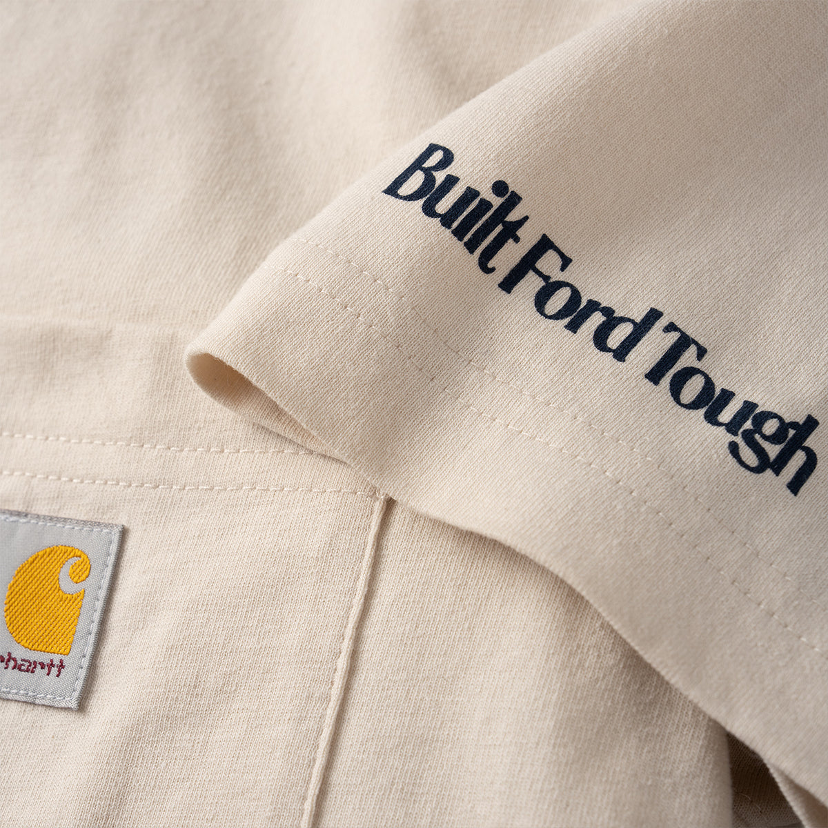 Close-up of a beige Carhartt work shirt showing the 'Built Ford Tough' embroidery on the sleeve and the signature yellow Carhartt logo patch on the chest pocket.