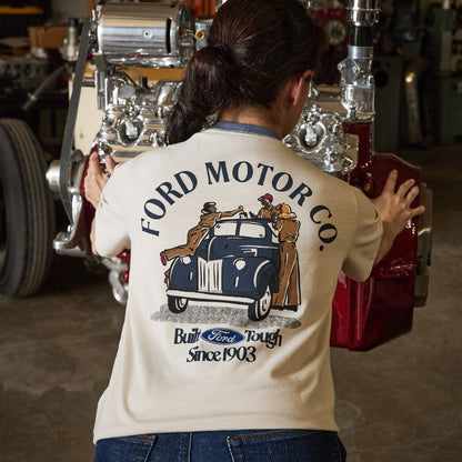 A woman viewed from behind wearing a cream-colored t-shirt with a vintage-style Ford Motor Co. graphic on the back, standing in a garage next to a large chrome engine and red car part.