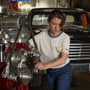 A woman in a Carhartt short-sleeve t-shirt and blue jeans works on a polished chrome engine in a garage with a classic car in the background.