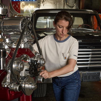 A woman in a Carhartt short-sleeve t-shirt and blue jeans works on a polished chrome engine in a garage with a classic car in the background.