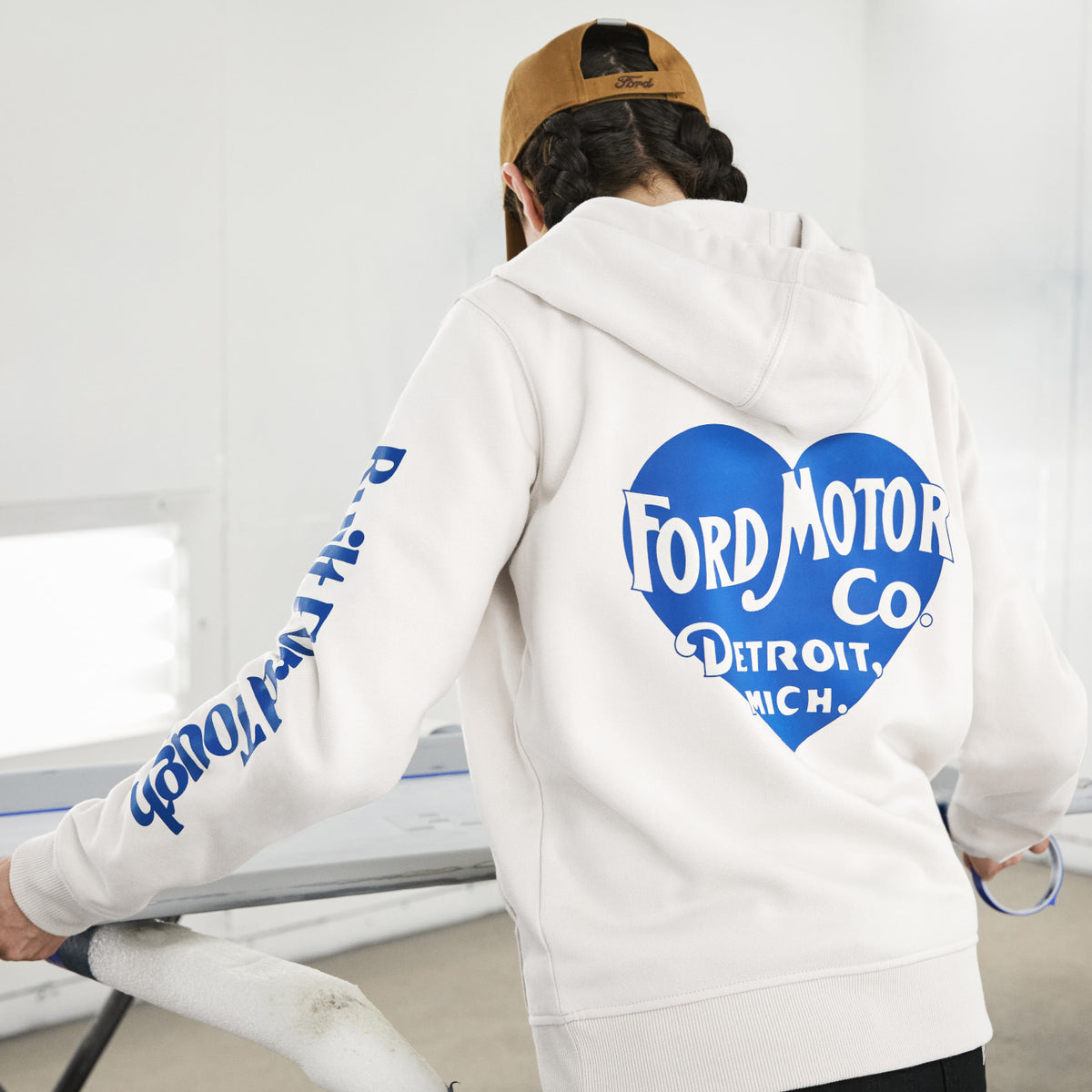 A person wearing a white hoodie with a large blue heart-shaped Ford Motor Co. graphic on the back and blue text down one sleeve, photographed from behind in a bright indoor setting.