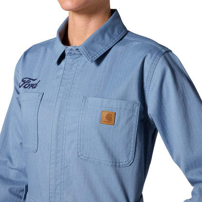 A close-up of a person wearing a light blue long-sleeve work shirt with a collar, two chest pockets, a dark embroidered Ford logo on the left chest, and a brown Carhartt brand patch on the right pocket.