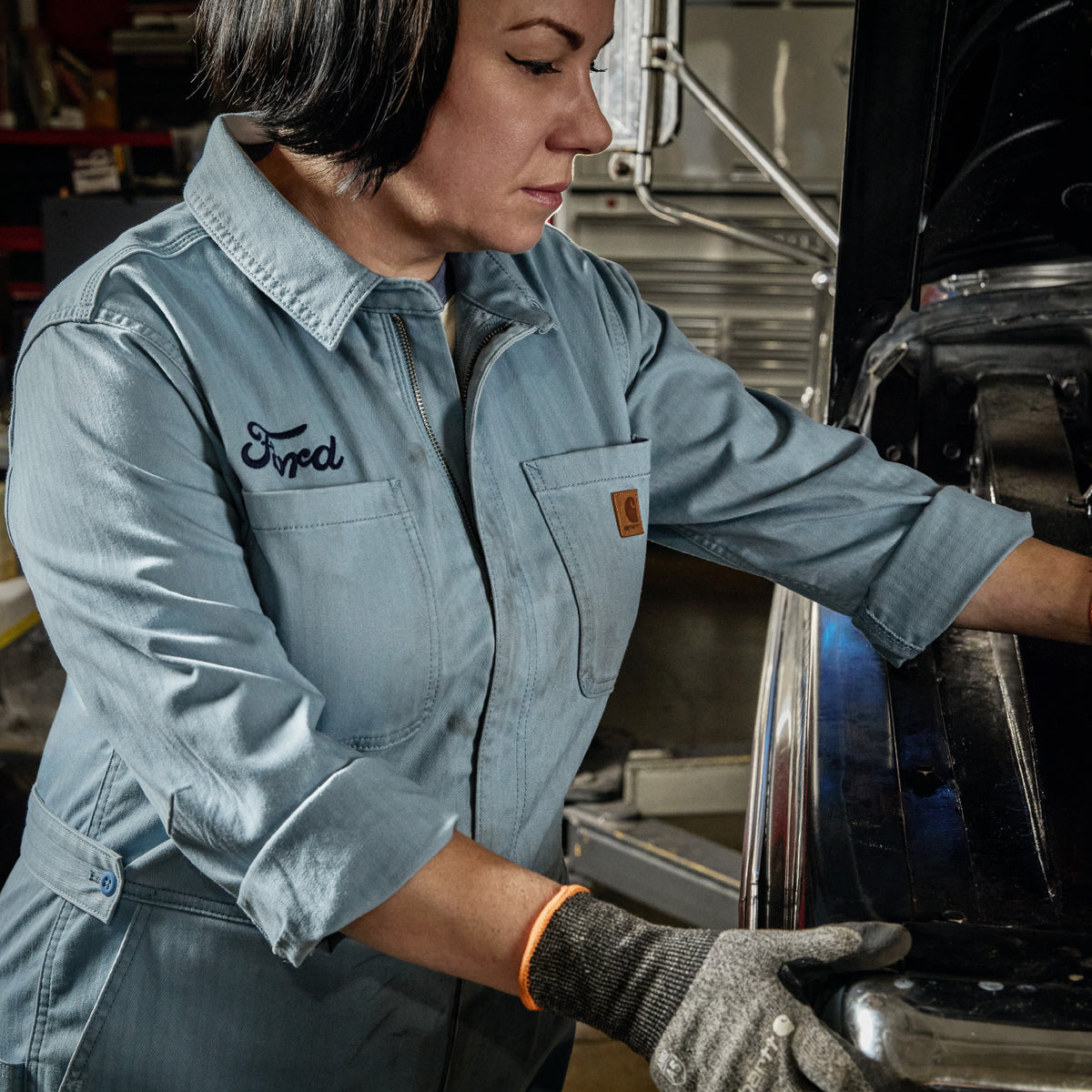 A woman in a light blue denim work shirt and gray work gloves works on a vehicle in a garage. The shirt has 'Fred' embroidered on the chest pocket.