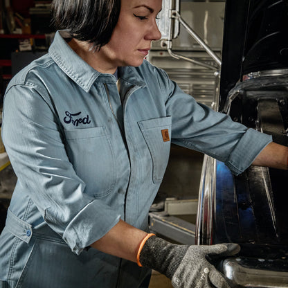 A woman in a light blue denim work shirt and gray work gloves works on a vehicle in a garage. The shirt has 'Fred' embroidered on the chest pocket.