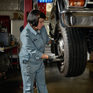 A woman in a light blue Ford jumpsuit and black gloves works on a car wheel in an auto repair shop.