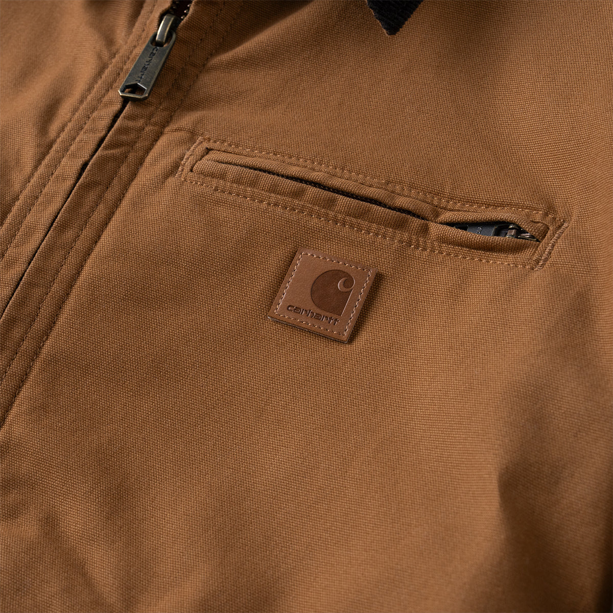 Close-up of a brown Carhartt jacket showing a zippered pocket with a brown leather Carhartt logo patch.