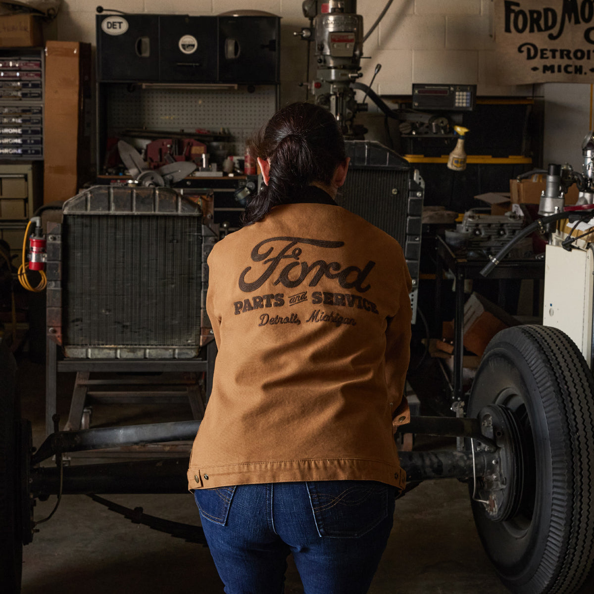 A person viewed from behind wearing a tan Ford Parts of Service jacket in a workshop, standing next to a vintage car frame with a large tire.