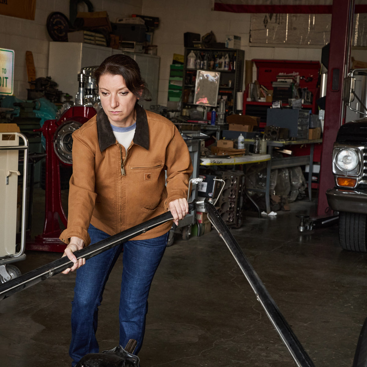 A woman in a brown Carhartt jacket and blue jeans stands in a cluttered garage holding a long metal bar.