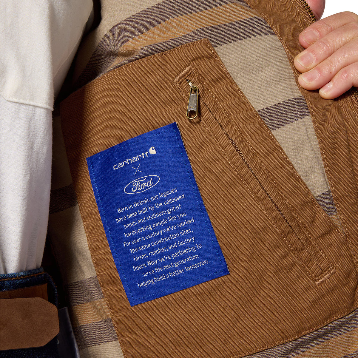 Close-up of a brown Carhartt x Ford patch pocket with a blue brand label, shown inside a plaid garment with a hand holding the fabric.