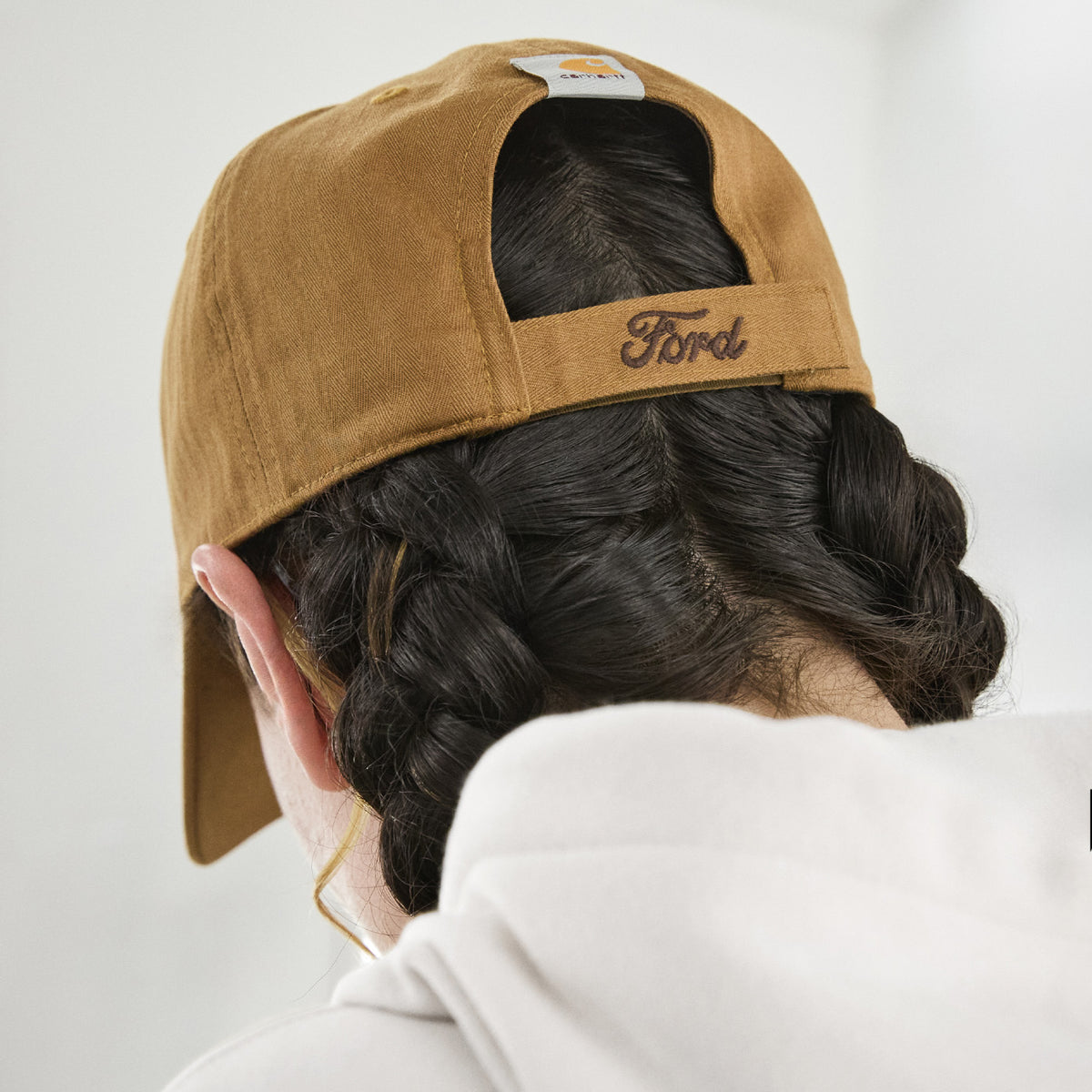 A person shown from behind wearing a tan Ford baseball cap and a white hoodie, with dark braided hair visible.