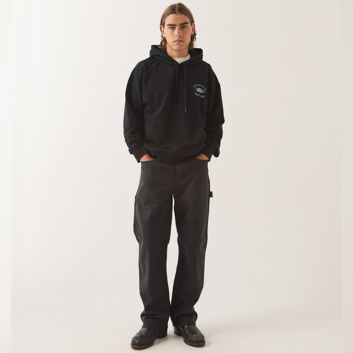 A model stands against a plain light background wearing a black pullover hoodie with a small blue and white graphic on the left chest, paired with dark charcoal cargo pants and black shoes.